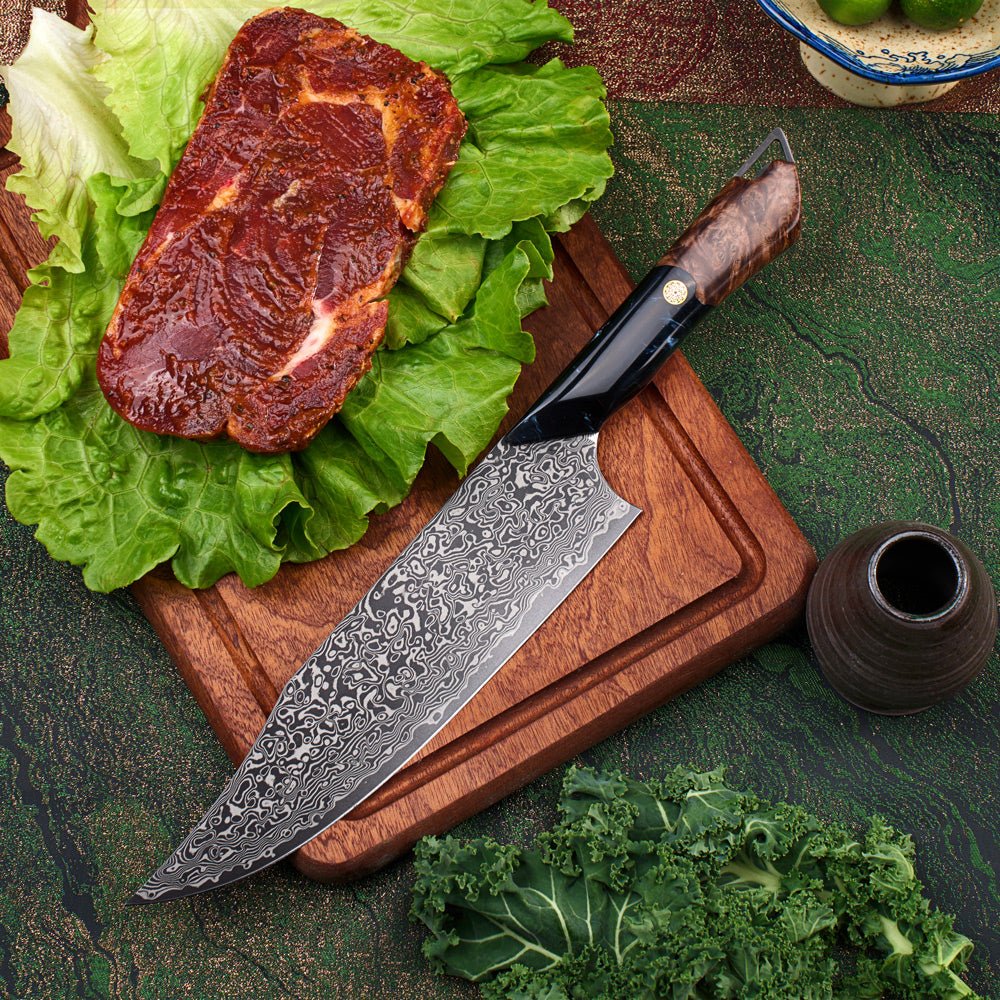 A Japanese-style chef's knife with a patterned Damascus steel blade and a brown wooden handle, resting on a wooden cutting board. The cutting board is styled with fresh green lettuce and a raw marbled steak, set against a dark green textured background with a small ceramic vase and leafy greens.