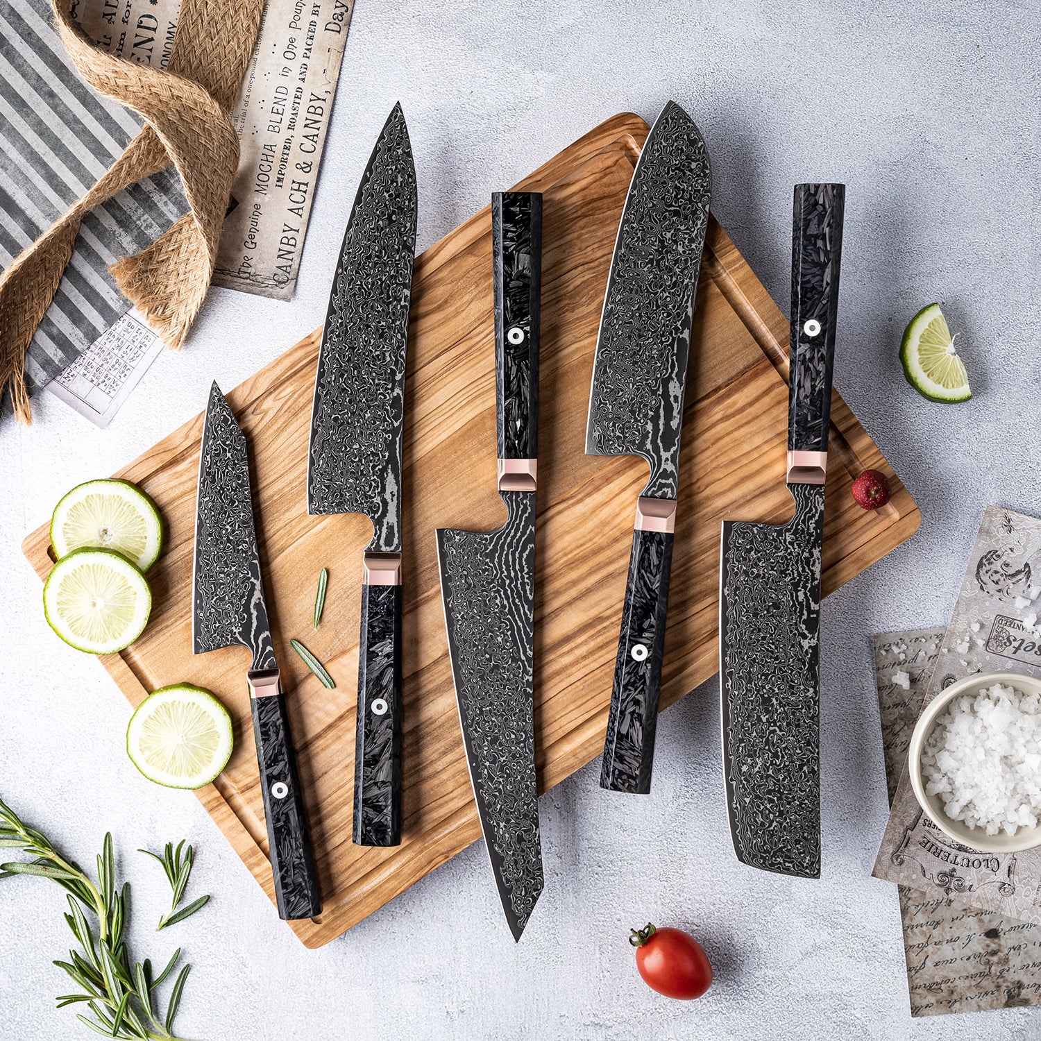 Set of Damascus steel kitchen knives on a wooden cutting board with limes and rosemary.