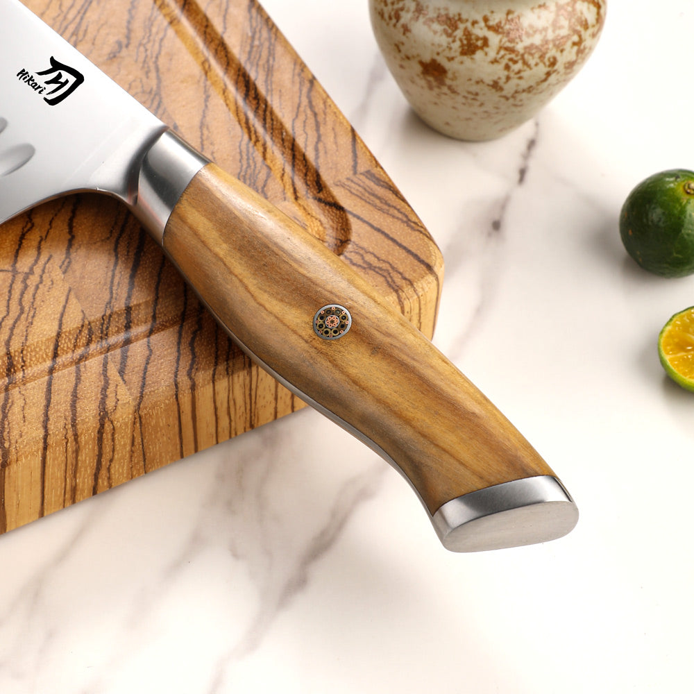 A chef's knife with a polished wood handle and silver metal bolster, resting diagonally on a matching wood cutting board over a white marble surface. A ceramic vase and sliced limes are visible in the background.