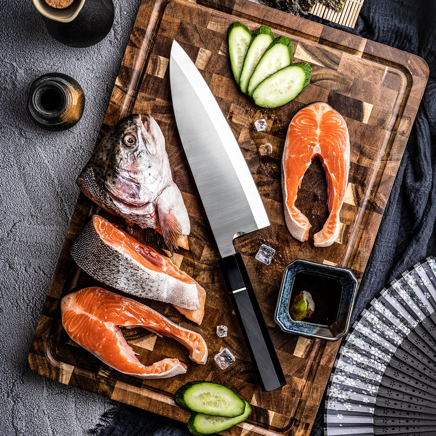 Hikari Knife Premium Deba knife for fish cuts on board, Raw fish fillets, and sliced cucumbers on a wooden cutting board with a dark background.
