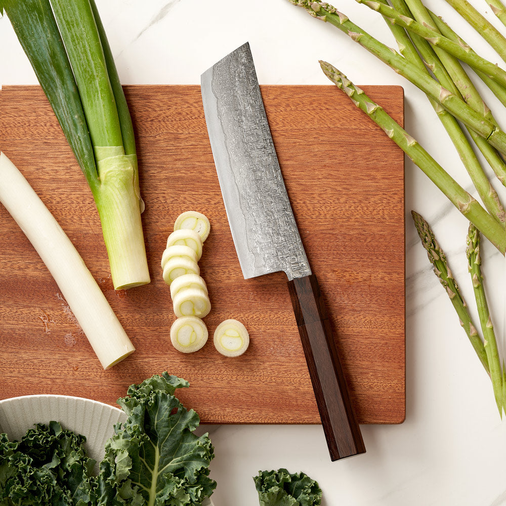 Wooden cutting board with sliced vegetables and a knife, surrounded by asparagus and leeks.