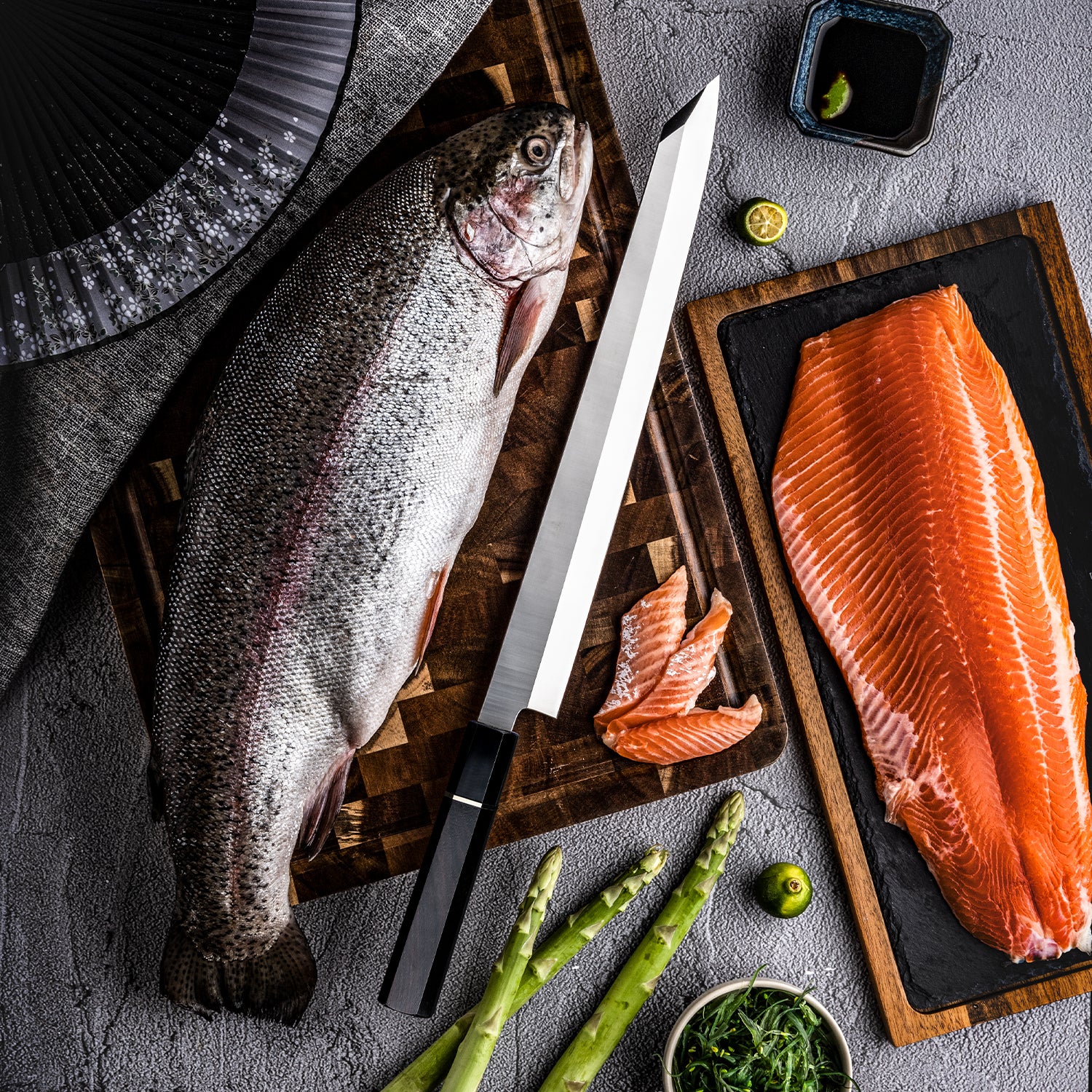 Premium Japanese fish knife with buffalo horn handle and carbon steel blade with Raw fish on a cutting board, lime, and asparagus on a gray surface.